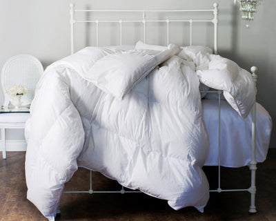 The St. Geneve Lajord Duvet, filled with 850 fill power Canadian Hutterite white goose down, is draped over a metal-frame bed in a minimal bedroom with a chair, table, and small floral arrangement in the background.
